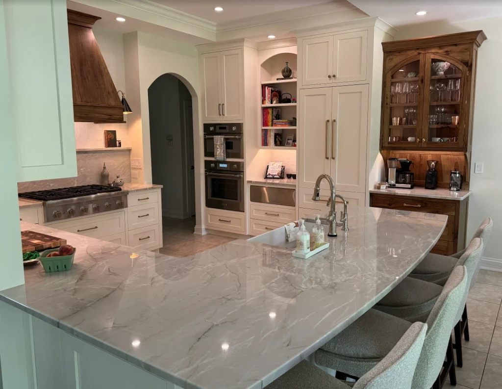 High-end kitchen featuring marble island with sink and upholstered bar stools, cream cabinetry, stainless steel gas range with wooden hood, double wall ovens, built-in refrigerator, and coffee station with glass cabinet.