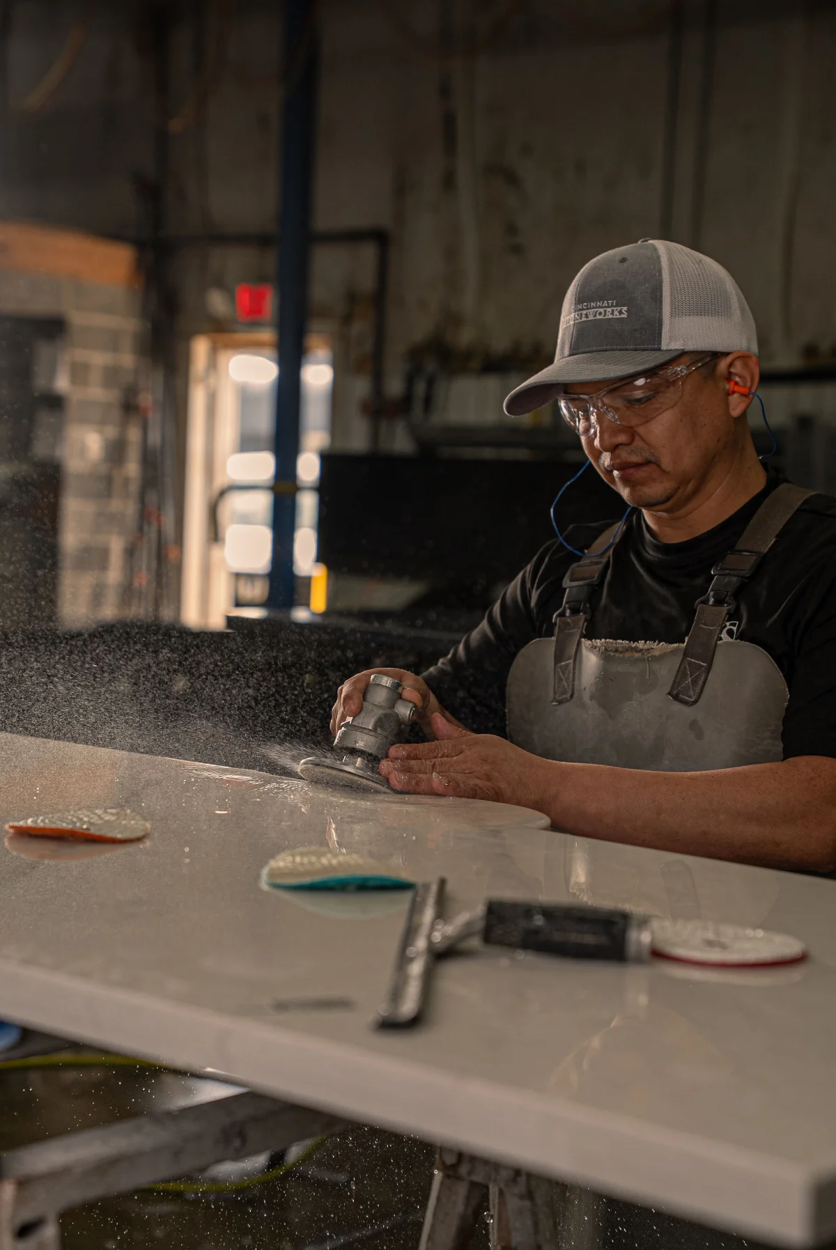Worker wearing protective gear sanding countertop surface with handheld power tool, fine particles spraying, polishing pads and squeegee on table, industrial workshop background.