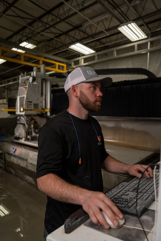 Factory worker operating computer workstation beside large industrial machine, wearing safety gear including hard hat and gloves.