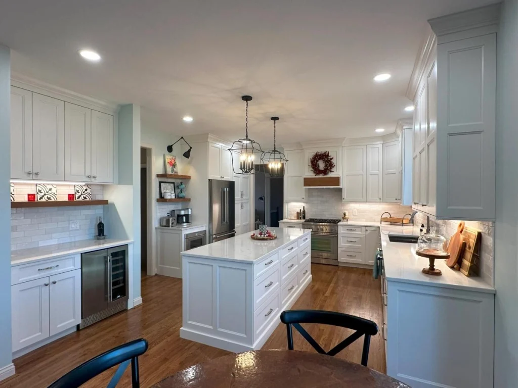 Modern kitchen featuring white cabinetry, marble island with drawers, stainless steel appliances including refrigerator, oven, microwave, and wine cooler, subway tile backsplash, pendant lights, warm wood flooring, and decorative red accents.