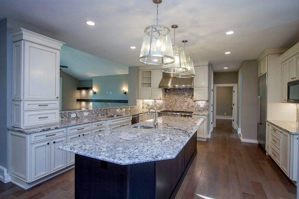 Elegant kitchen featuring large granite island with sink, white cabinetry with dark hardware, stainless steel appliances, clear glass pendant lights, and dark wood flooring.