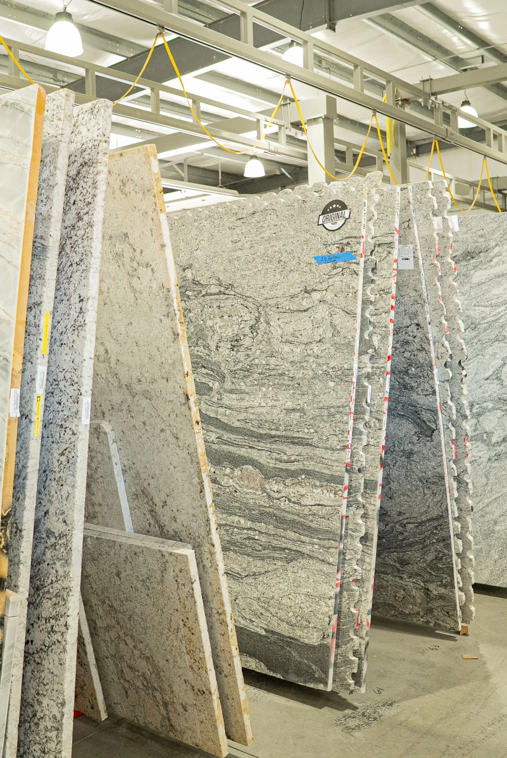 “Warehouse showroom with large granite and marble slabs standing vertically in rows, each labeled with tags for identification under industrial lighting.”