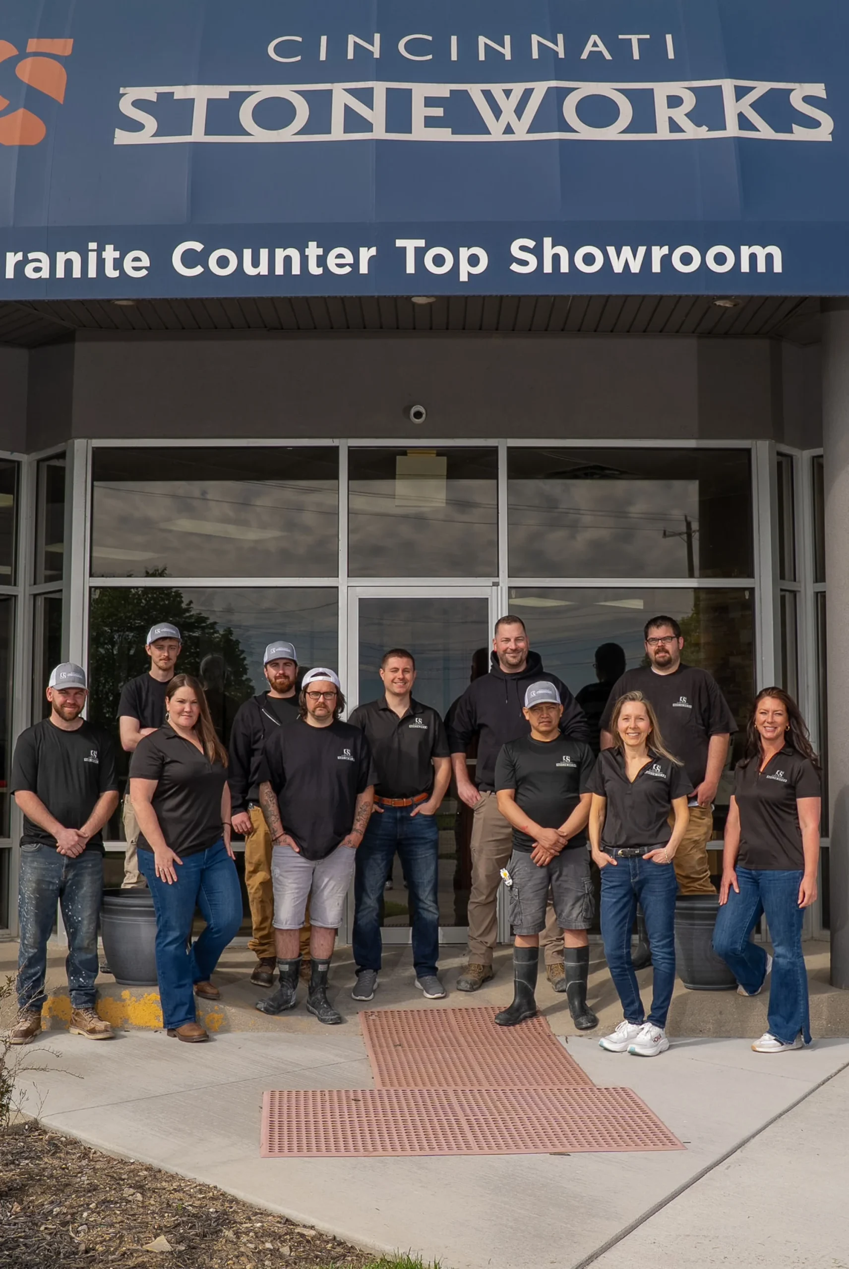 Group of eleven staff members standing in front of Cincinnati Stoneworks granite countertop showroom, under large blue awning with business name and description.
